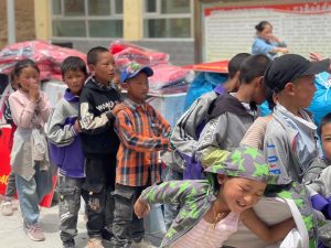 Niños en fila con uniformes de trabajo de varios colores frente a un edificio con suministros.