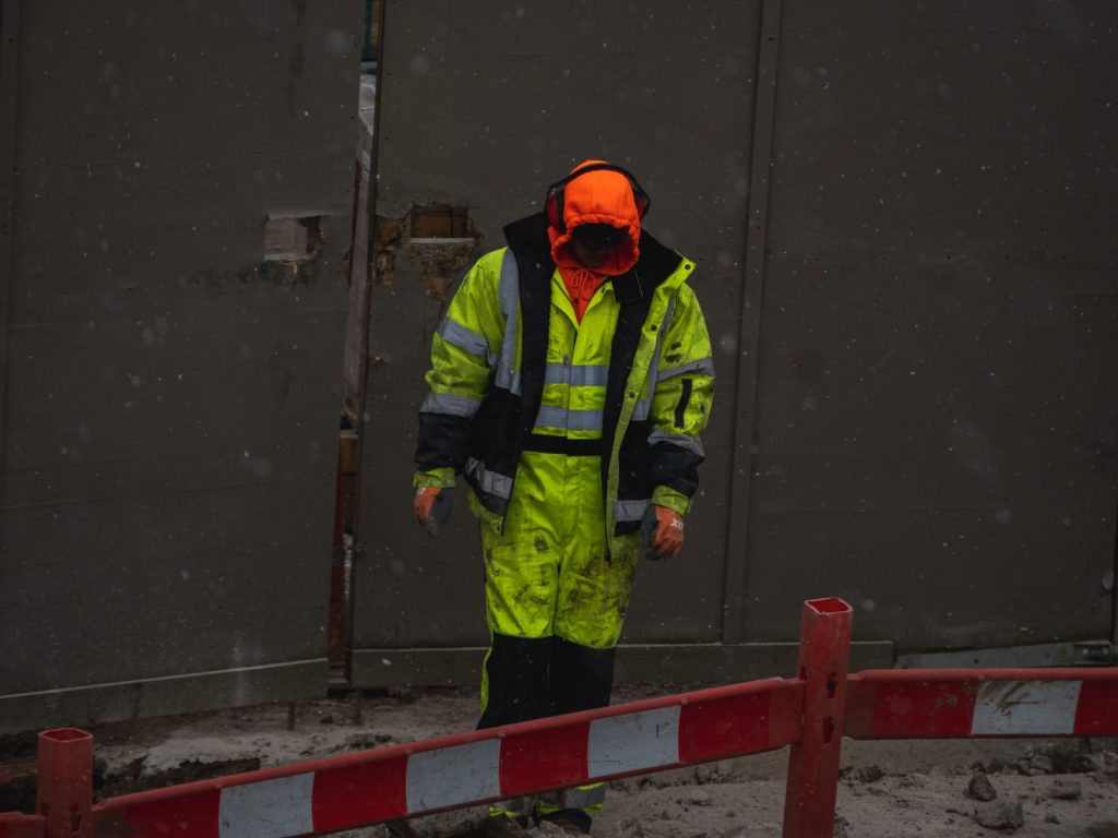 High-visibility construction worker wearing reflective safety gear, waterproof coat, and protective gloves standing on a construction site in rainy weather.