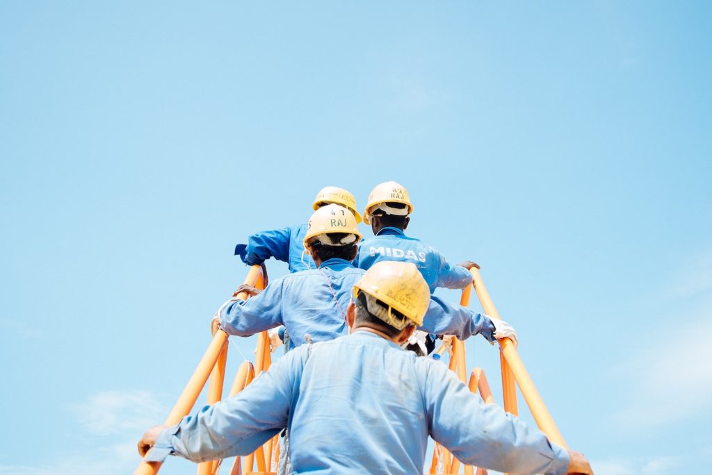 Workers in blue uniforms and yellow safety helmets climbing an orange ladder against a clear blue sky.