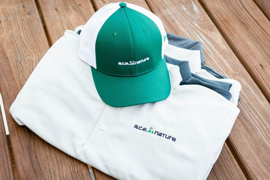 Green and white baseball cap with custom logo on front and mesh back, placed on top of folded white and gray company uniforms, on wooden floor.
