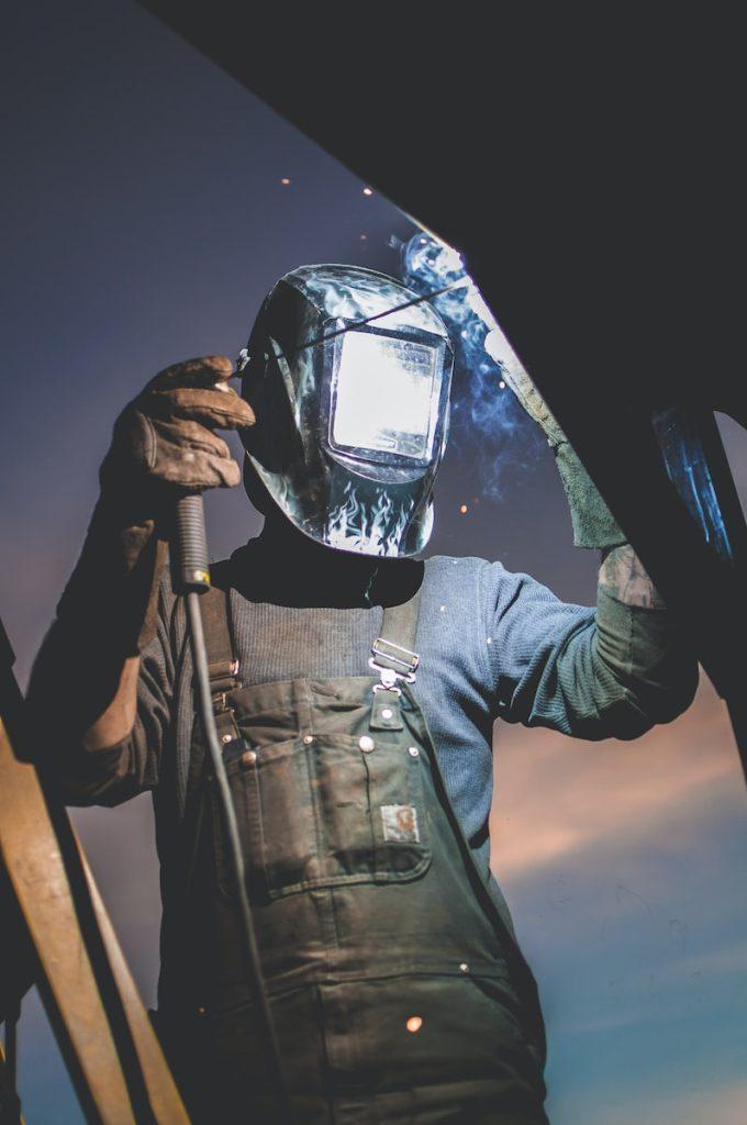 Welding worker wearing protective helmet and gloves welding metal outdoors at sunset.