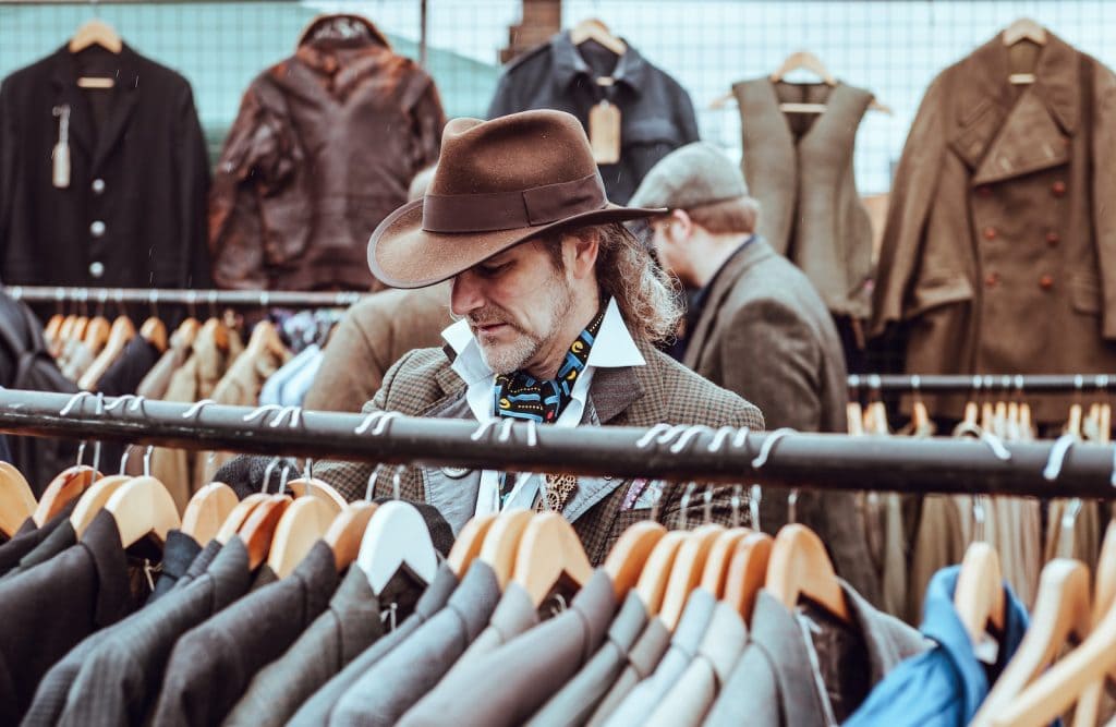 Stylish man shopping for vintage or custom clothing jackets at a retail store, focused on trendy fashion selections with a variety of jackets hanging on racks behind him.
