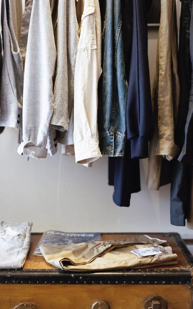 Assorted casual and workwear shirts hanging on a clothing rack with folded pants on a wooden table underneath.