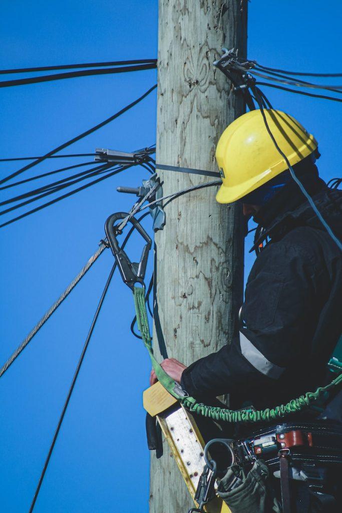High-voltage electrical worker repairing power lines on utility pole, wearing safety helmet and workwear, performing maintenance work on electrical infrastructure.