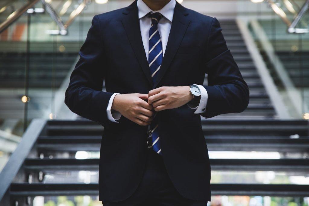 Polished businessman in a tailored dark suit, white shirt, and striped tie standing on modern stairway in an office building.