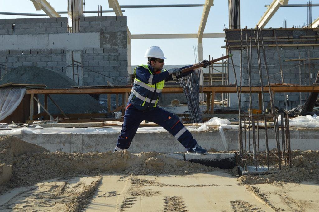 Workwear construction worker wearing safety vest and helmet at a building site, handling rebar in a concrete construction project.