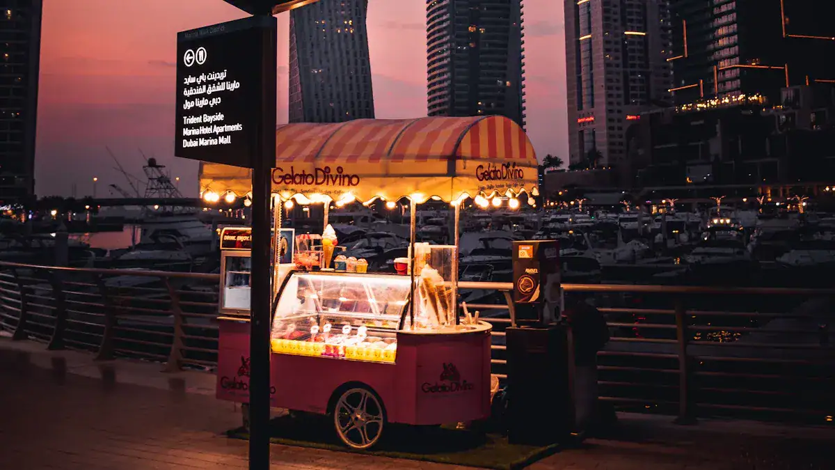 Vibrant pink and yellow ice cream cart selling treats at dusk along a waterfront with modern high-rise buildings in the background.