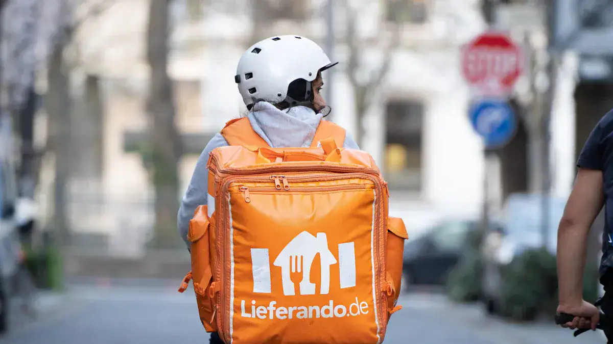 Delivery person wearing an orange insulated backpack with Lieferando.de logo for food delivery on city street in Germany.