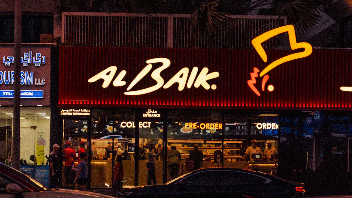 Al-baik restaurant illuminated sign in vibrant red and yellow colors at night; busy storefront with customers inside and outside; prominent logo with hat illustration; inviting fast-food ambiance.