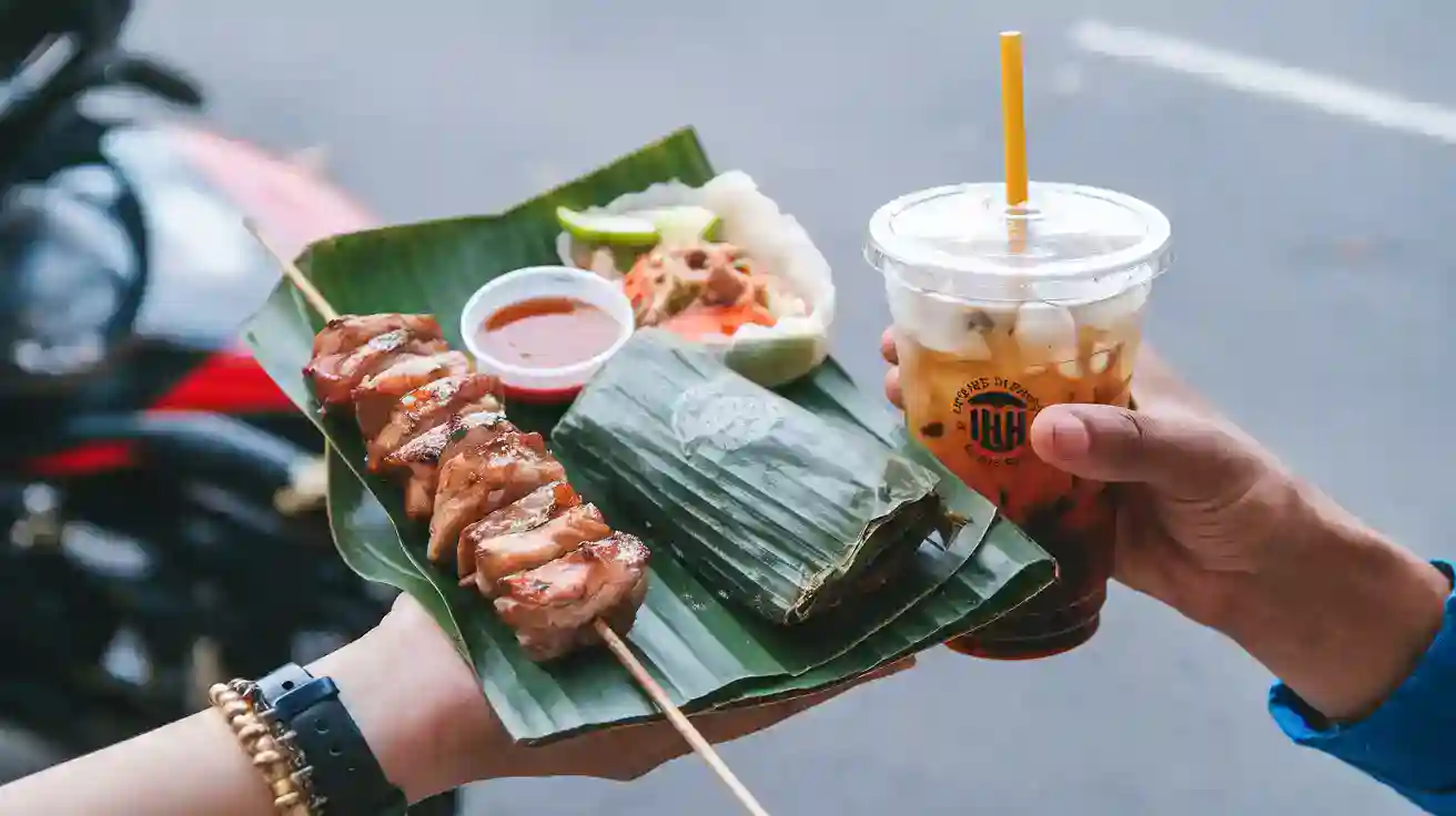 Sweet and savory grilled chicken skewer, traditional Vietnamese spring roll, and iced tea served on banana leaf in street food setting.