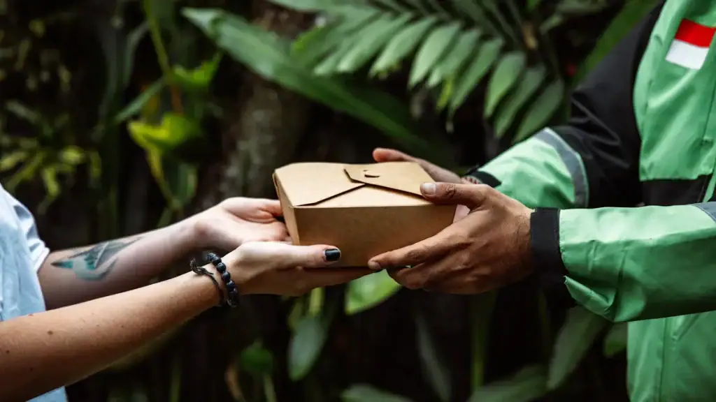 Delivery person handing over a package to a customer in an outdoor setting with lush green foliage in the background, showcasing eco-friendly packaging and customer service excellence.