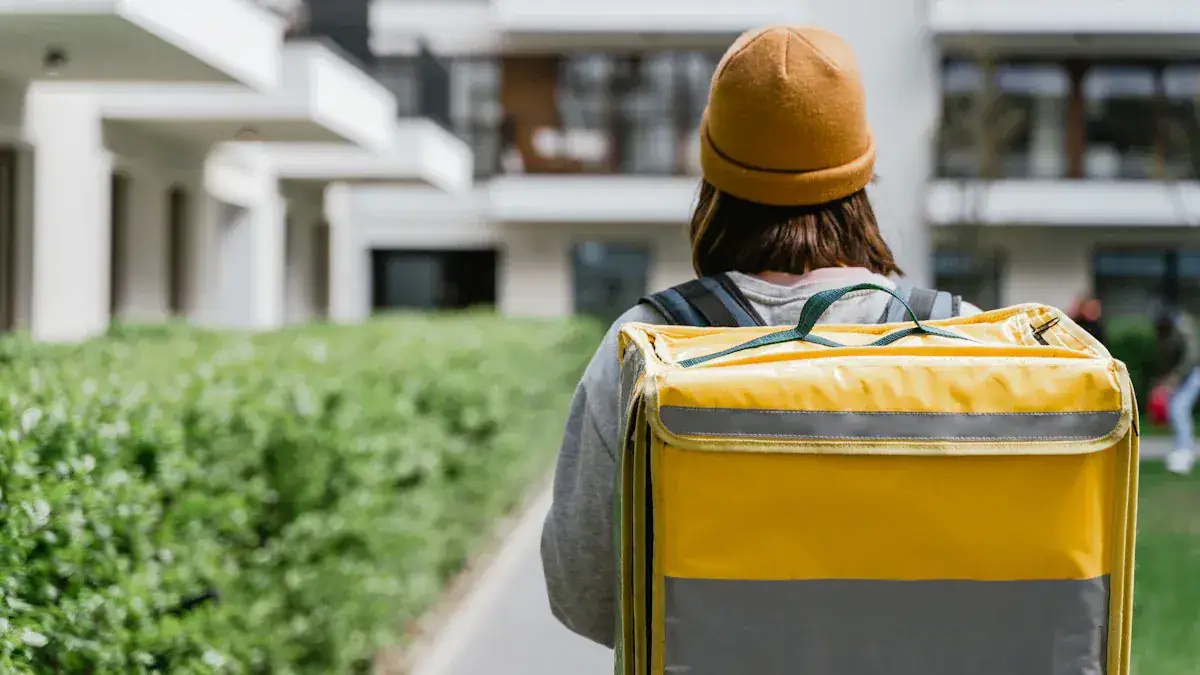 Delivery person carrying a yellow insulated food delivery bag, wearing casual clothing and a brown beanie, outside modern apartment buildings, ready for food delivery service.