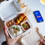 Freshly prepared Asian-style takeout in eco-friendly packaging with rice, skewers, and dipping sauces on a white table. Smartphone nearby, ready to enjoy a delicious meal.