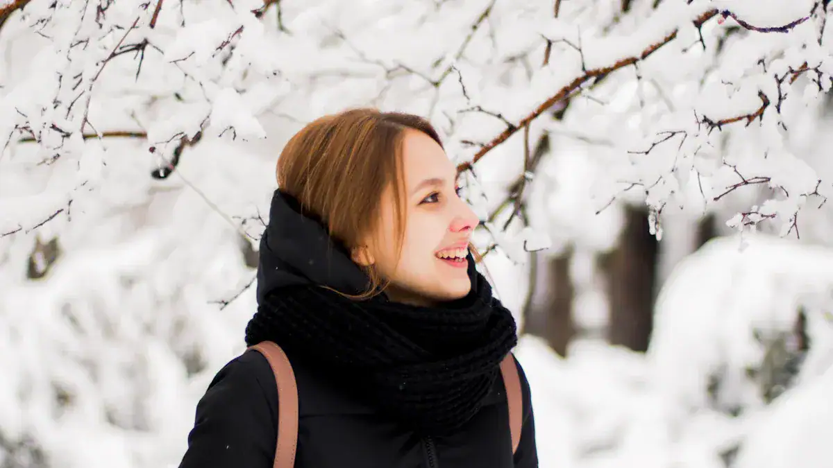 Woman with brown hair smiling in winter snow scene, wearing warm black jacket and scarf, enjoying outdoor snowy environment.