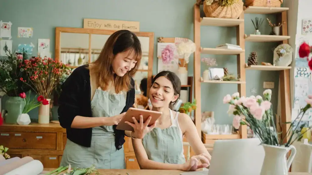 Flower shop workers discussing floral arrangements, floral shop interior, happy women in aprons, floral display shelves, professional floral artistry, flower shop environment, custom floral clothing uniforms China manufacturer.