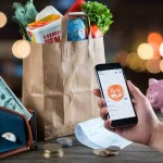 Reusable shopping bag with groceries and cash on table, person making digital payment via smartphone.