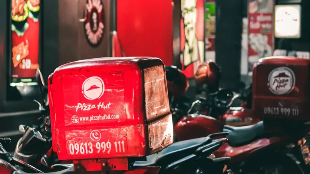 Red Pizza Hut delivery scooter with company logo and contact number parked among other vehicles in an urban street scene.