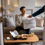 Fresh food delivery person handing over a meal to a smiling woman sitting on a cozy sofa in a modern living room. The scene captures a friendly food delivery experience.