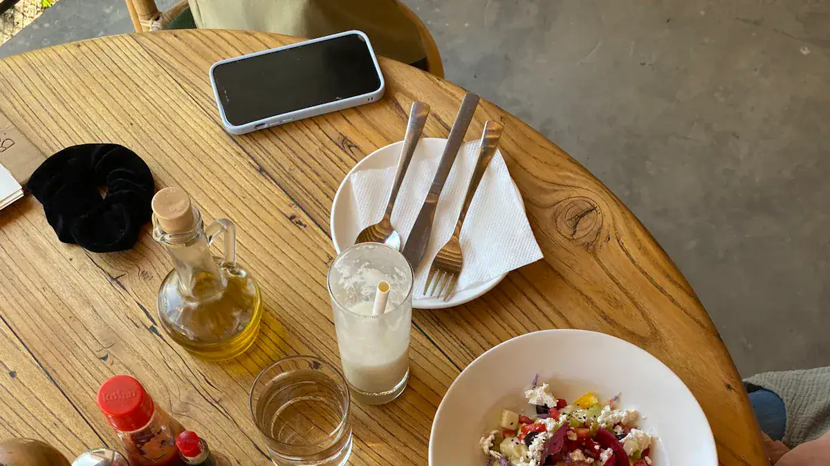 Fresh salad on white bowl with cutlery and drink on wooden table in casual dining setting, top view of restaurant food presentation and table setup, healthy meal with beverages and utensils, visually appealing cuisine scene for restaurant promotion.