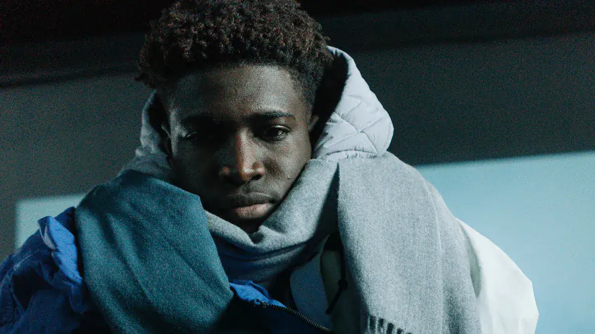 Young man in casual winter clothing, including a hoodie, jacket, and scarf, looking pensive in a dark indoor setting.