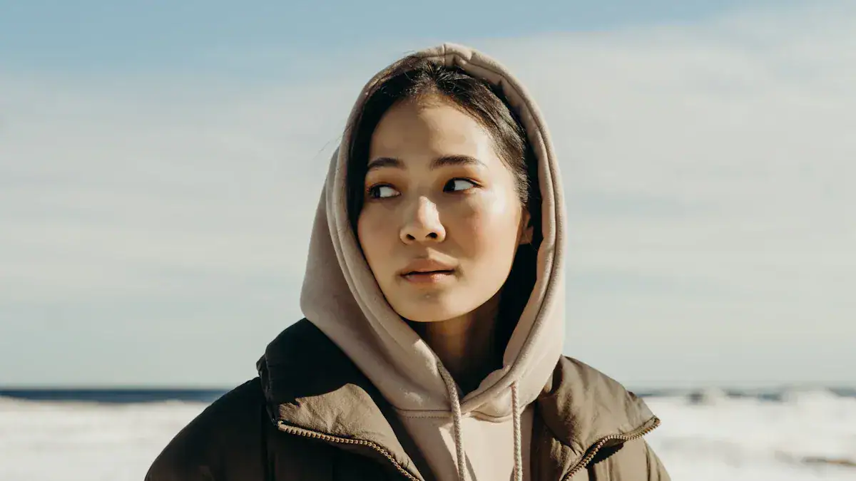 A woman in a hoodie and jacket standing outdoors near the beach on a cloudy day.
