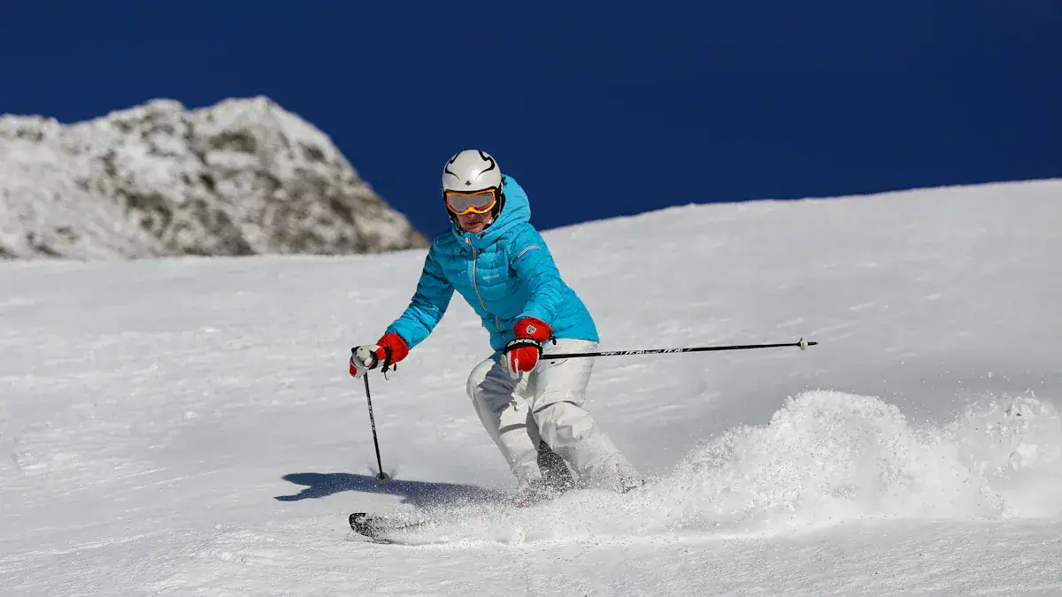 Bright blue ski jacket, white ski pants, helmet, and goggles worn by a skier descending snowy mountain slope on a sunny day.