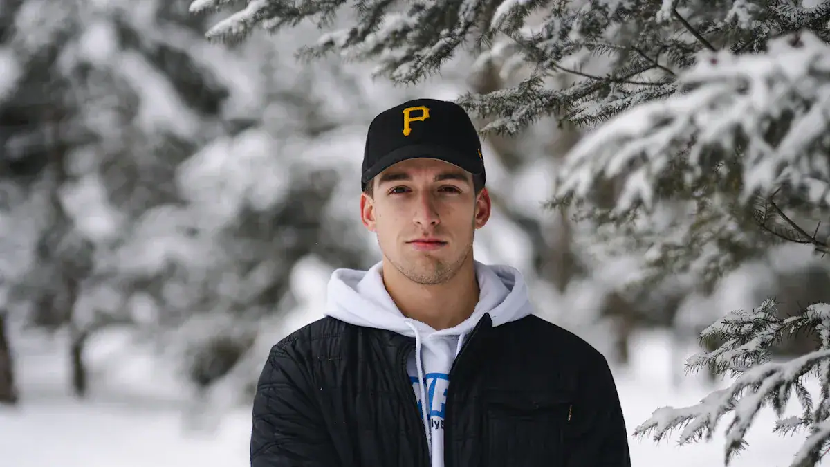 Warm winter clothing in a snowy outdoor setting, featuring a young man wearing a black cap with a yellow team logo, a white hoodie, and a black jacket. Perfect for cold-weather workwear and outdoor activities.