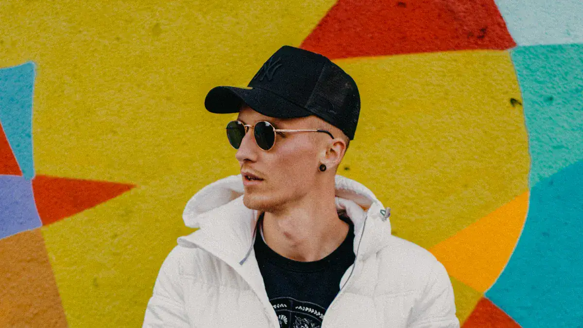 Black baseball cap, sunglasses, and white hooded jacket model standing against colorful graffiti wall. Stylish casual fashion with custom clothing options from China manufacturer.