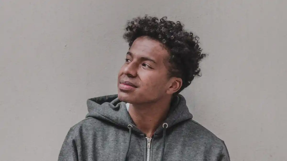 Comfortable young man in casual hoodie, gray sweatshirt, and textured afro hairstyle, standing against a plain wall for a relaxed and stylish look.