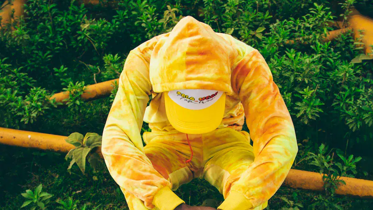 Colorful tie-dye hoodie and pants set with a yellow cap, designed for casual or outdoor wear, worn by a person crouching in a lush green environment.