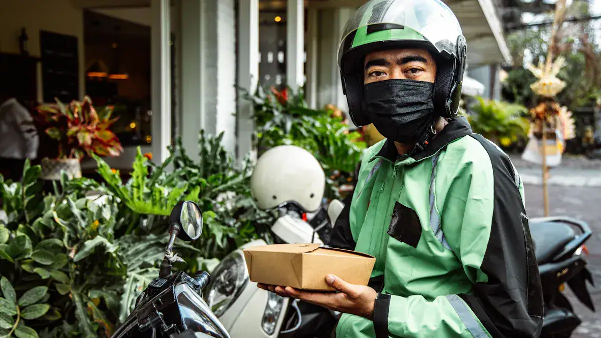 Delivery person wearing a black mask and helmet, holding a brown food package next to scooters and greenery.