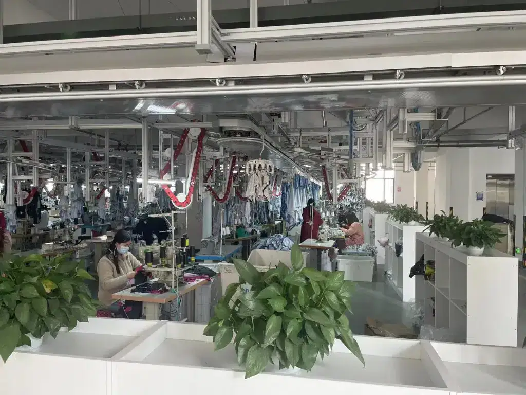 White factory interior with workers sewing and assembling uniforms in a modern garment manufacturing plant green plants in the foreground and workplace automation equipment in the background alt text: modern garment manufacturing factory with workers sewing uniforms and green indoor plants caption: a spacious garment factory in China specializing in custom clothing uniforms with active workers and contemporary machinery image description: this image showcases a well-organized Chinese clothing manufacturing factory where workers are engaged in sewing and assembling custom uniforms. The factory features modern equipment, greenery for a pleasant environment, and an efficient workflow, emphasizing quality apparel production relevance: high.
