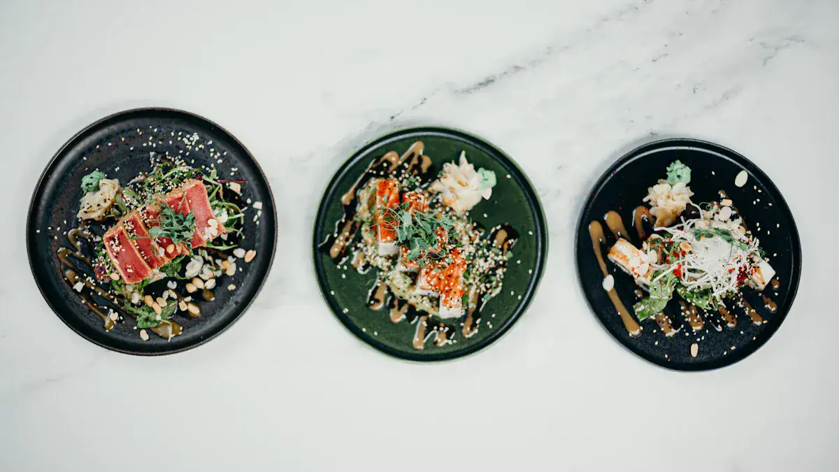 Seared tuna sashimi, sushi rolls, and fresh salad on black ceramic plates, garnished with sesame seeds and greens, served on a white marble surface.