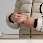Elegant beige puffer jacket with black cuffs, paired with a modern white table decorated with sleek gold and black pens, creating a sophisticated workspace aesthetic.