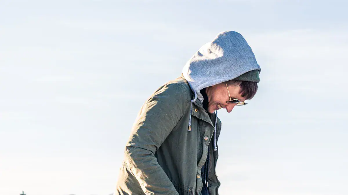 Brightly dressed man wearing layered casual outdoor clothing, including a hoodie and jacket, smiling while looking down against a clear sky background.