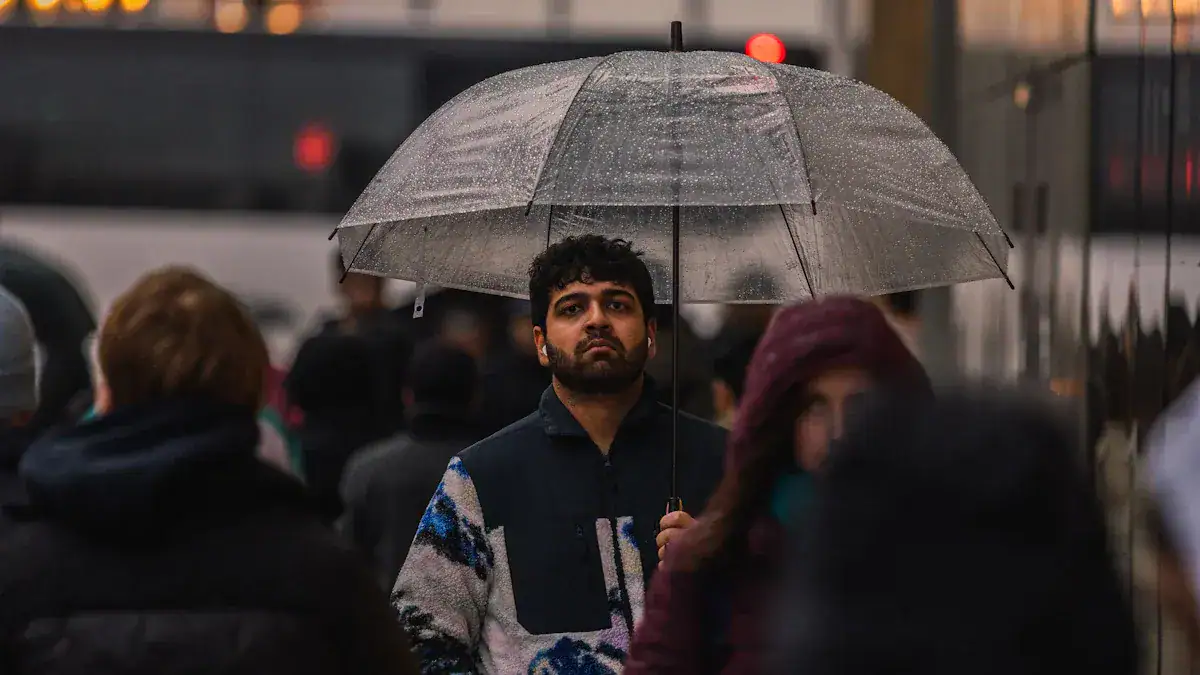 Rainy day urban street scene with man holding transparent umbrella among crowd of people in weather-appropriate clothing during drizzle or rain.