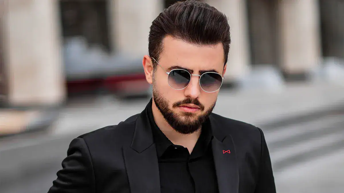 Sunglasses and stylish blazer worn by a man with a beard on city street.