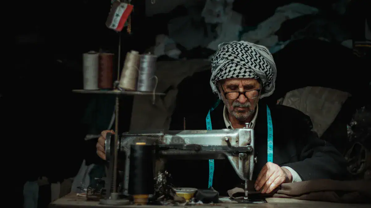 Old man sewing fabric with a vintage sewing machine in a workshop or tailor shop, surrounded by spools of thread and fabric materials.