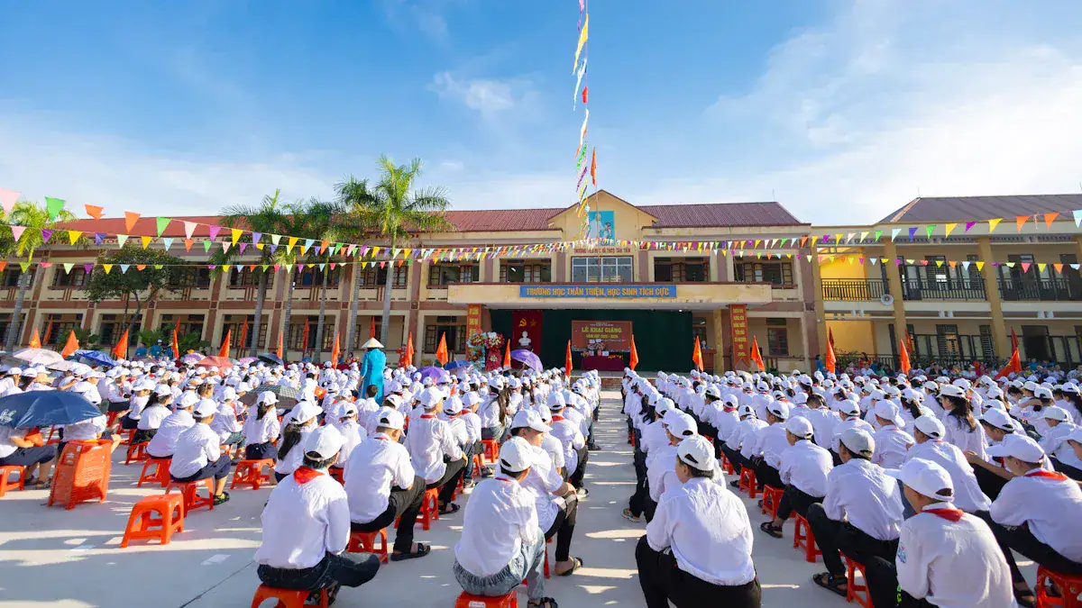 People wearing white uniforms and hats attending a school assembly outdoors with flags and banners in the background.