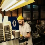 Stacked disposable coffee cups at a coffee shop counter with barista preparing drinks.