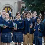 Students in school uniforms outside a school building, smiling and holding books, wearing custom-made blue blazers and plaid skirts or pants, representing educational uniforms from a Chinese manufacturer.