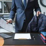 Elegant businessman holding a navy plaid blazer and a document in an office setting, showcasing custom clothing and uniforms manufacturing services by Sha.