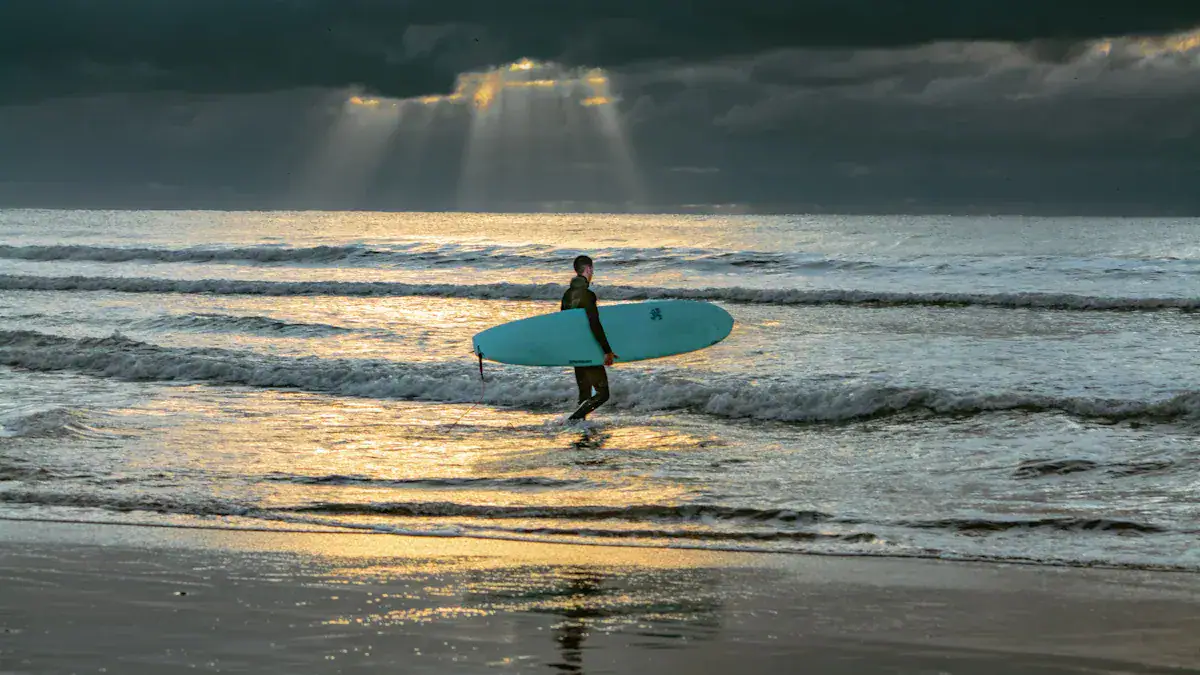 A surfer walking into the ocean at sunset, holding a blue surfboard with dramatic cloudy skies overhead.