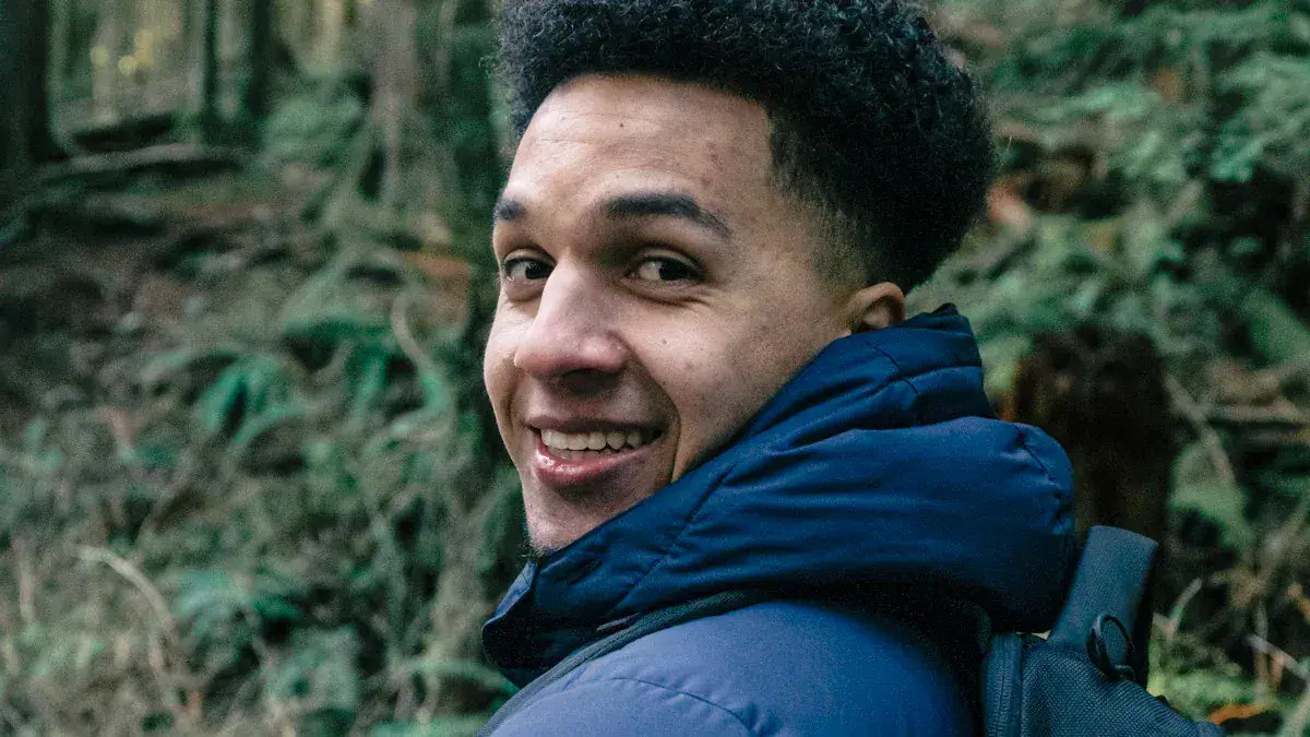 Men smiling outdoors wearing a blue jacket and backpack in a forest setting.