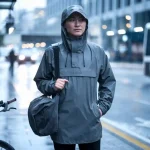 Waterproof gray rain jacket worn by a man with a backpack on a city street in rainy weather.