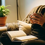 A person wearing a black sweatshirt sitting on a beige sofa, holding a mug with both hands while reading a book, with a potted plant on a small white table in the background.