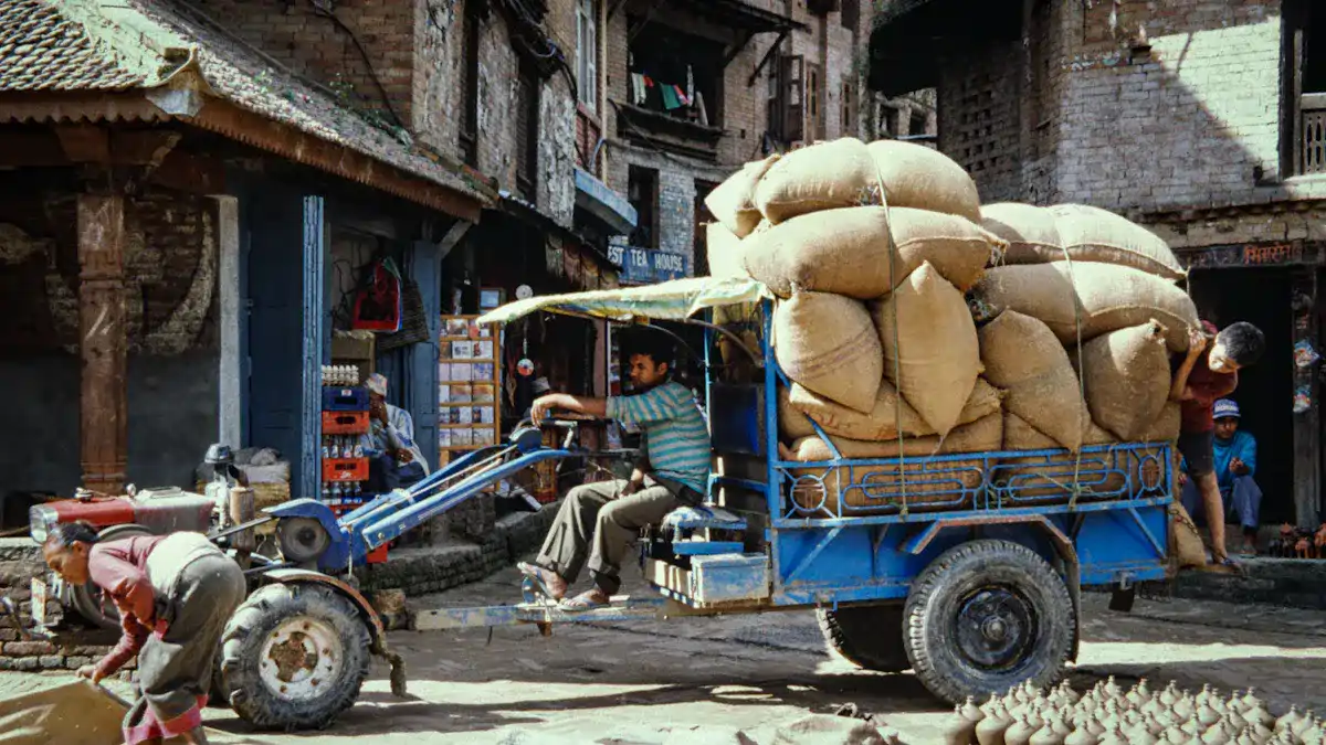 Colorful street scene featuring an old vehicle loaded with large sacks, surrounded by local residents and traditional brick buildings in a bustling village area.