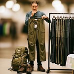 Olive green outdoor cargo pants displayed in a store, with hiking boots, a backpack, and a map on the table, emphasizing custom workwear for outdoor activities and adventure garments.