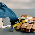 Folded blue denim jeans with fabric swatches labeled "DENIM," "CANVAS," and "TWILL," a yellow measuring tape, and a pair of protective work gloves, illustrating custom clothing and workwear manufacturing.